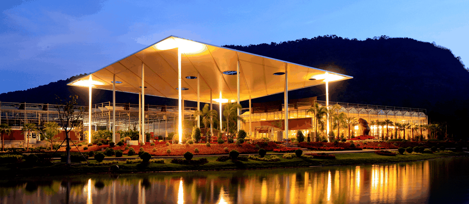 Large modern pavilion with an angular roof illuminated at dusk, overlooking a reflective pond with a mountain backdrop